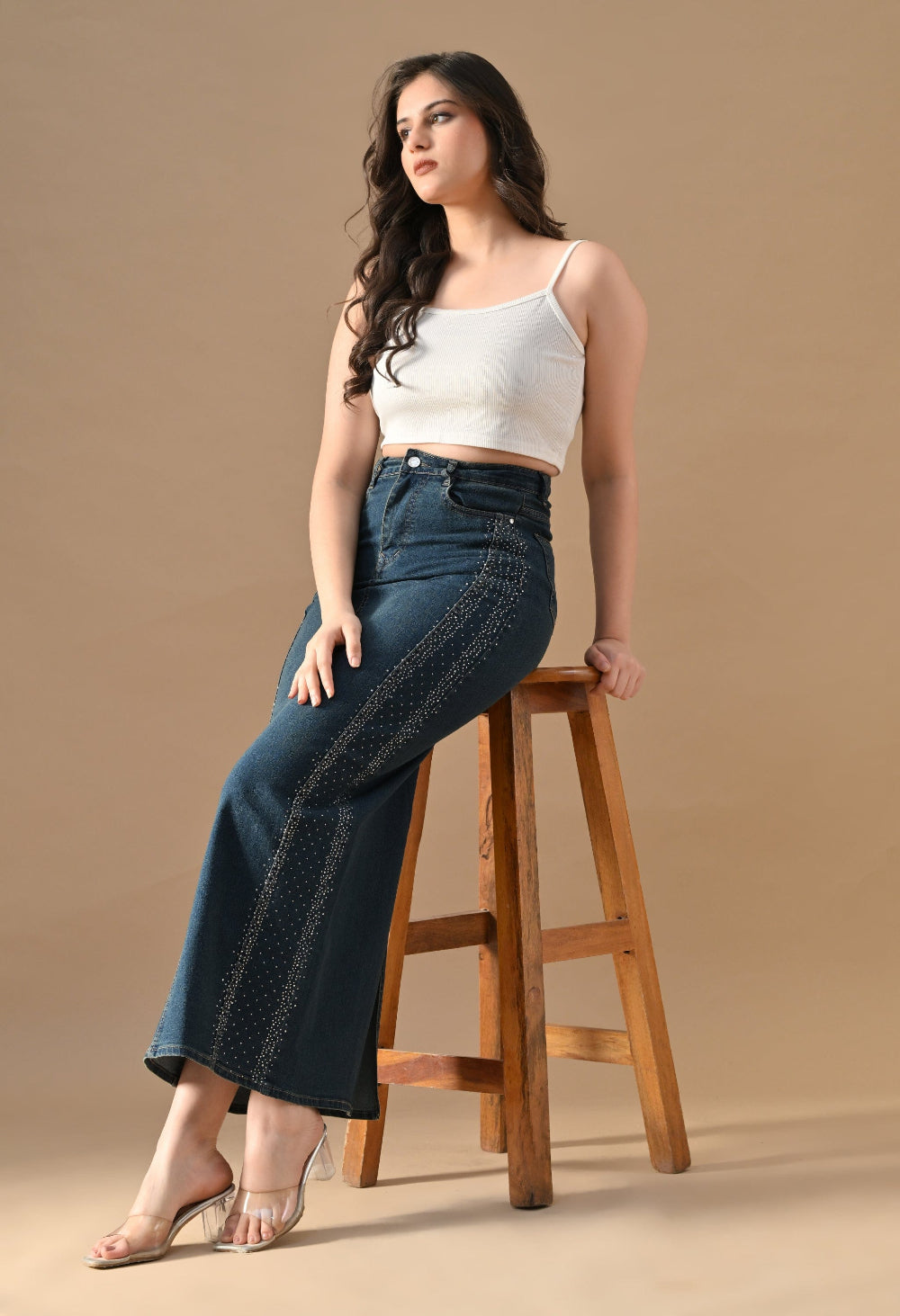 Woman wearing a white top and blue jeans sitting on a wooden stool against a beige background