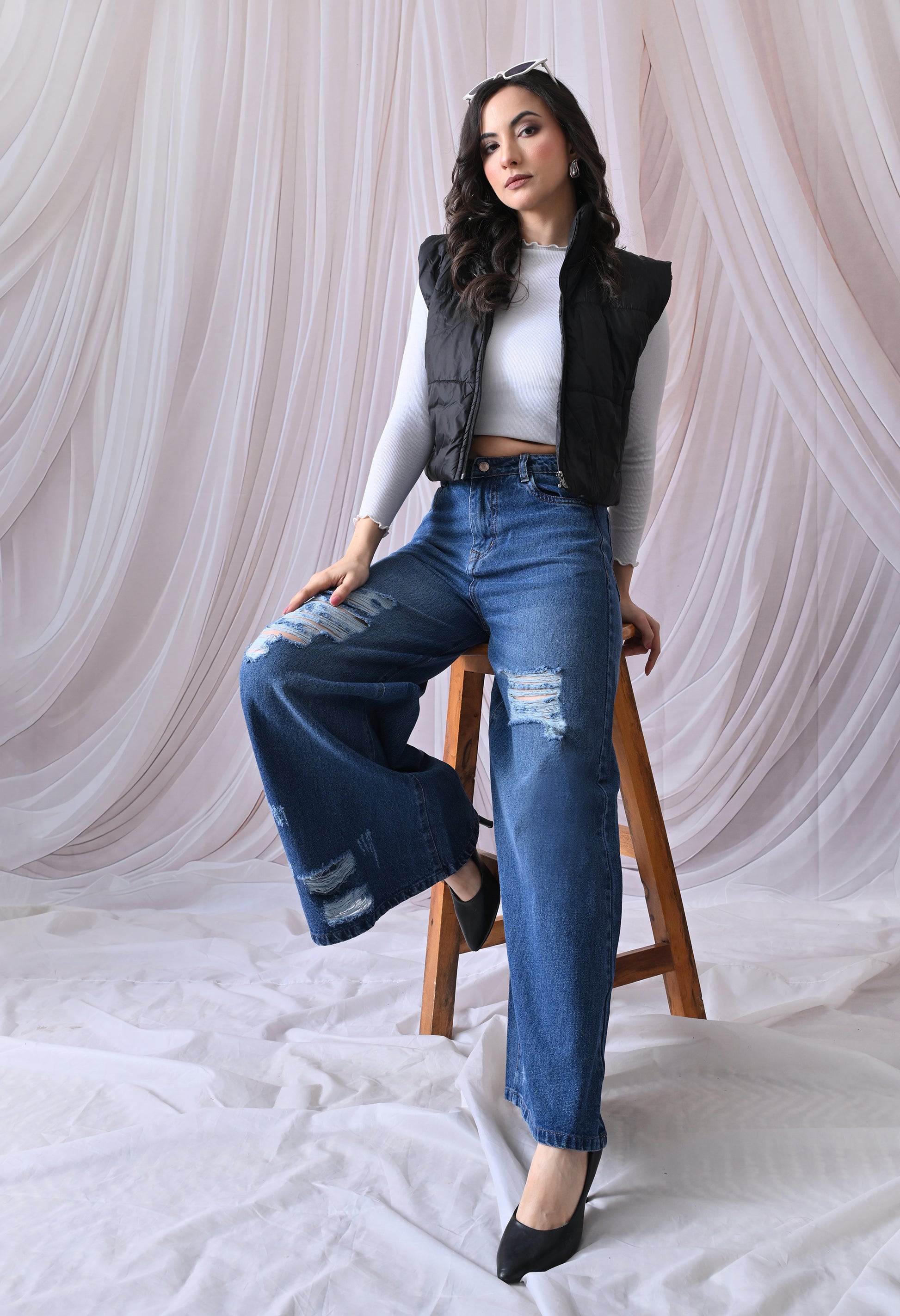 Woman sitting on a stool wearing a black vest, white top, and blue jeans against a white curtain background.
