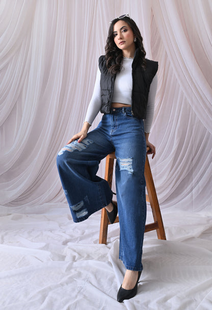 Woman sitting on a stool wearing a black vest, white top, and blue jeans against a white curtain background.