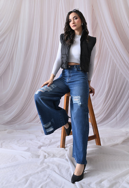 Woman sitting on a stool wearing a black vest, white top, and blue jeans against a white curtain background.