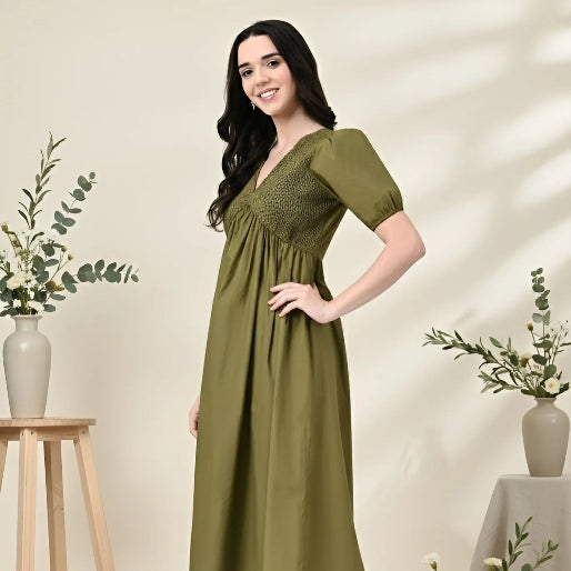 Woman wearing a green dress standing in a decorated room with plants and a stool.